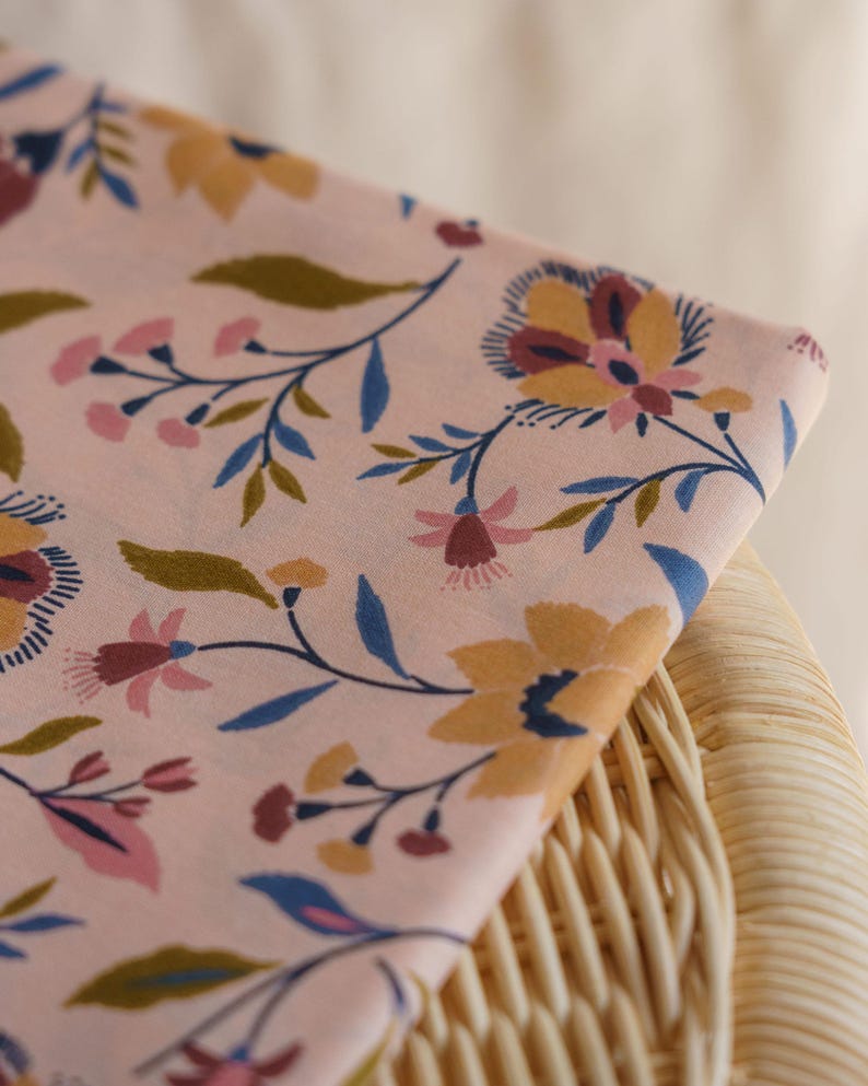 May include: A folded piece of light pink fabric featuring a repeating floral pattern. The design includes stylized flowers and leaves in shades of mustard yellow, dark blue, and muted pink, arranged across the soft-toned background surface.