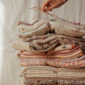 Op de afbeelding: Een stapel gevouwen stoffen die met touw aan elkaar zijn gebonden. De textielsoorten hebben diverse patronen, waaronder kleine bloemenprints, effen gedempte tinten en subtiele geometrische ontwerpen in crème, beige, perzik en zachte bruintinten.