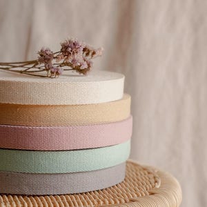 Puede incluir: Una pila de seis carretes de cinta tejida en varios colores pastel: crema, melocotón, rosa, verde menta y gris. Flores de lavanda secas descansan encima. Los carretes están apilados sobre un taburete de mimbre.