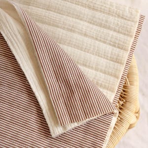 May include: A reversible quilted fabric piece featuring a cream-colored side with horizontal stitching and a brown and white pinstriped side. The textile has a soft, textured appearance with visible raw edges and a layered, folded construction.