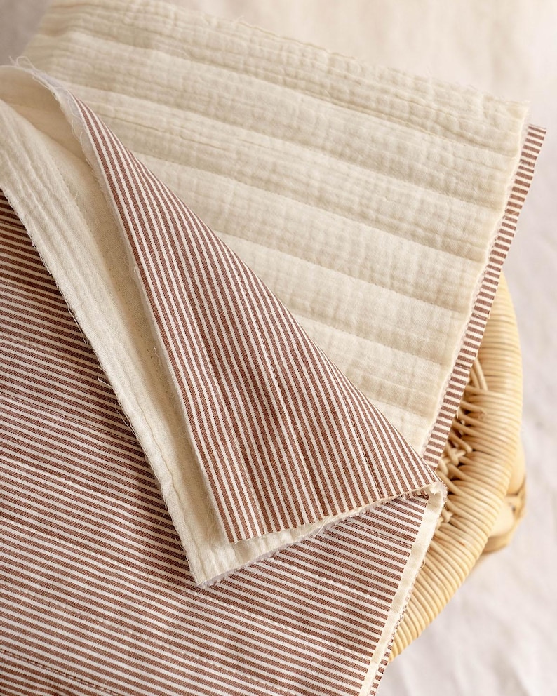 May include: A reversible quilted fabric piece featuring a cream-colored side with horizontal stitching and a brown and white pinstriped side. The textile has a soft, textured appearance with visible raw edges and a layered, folded construction.