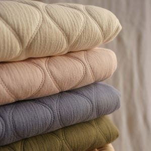 Può includere: Una pila di quattro coperte trapuntate in vari colori: crema, rosa cipria, blu ardesia e verde oliva. Ogni coperta presenta un motivo circolare cucito e ripetuto. Le coperte sono impilate, mostrando la loro morbida consistenza e il design.