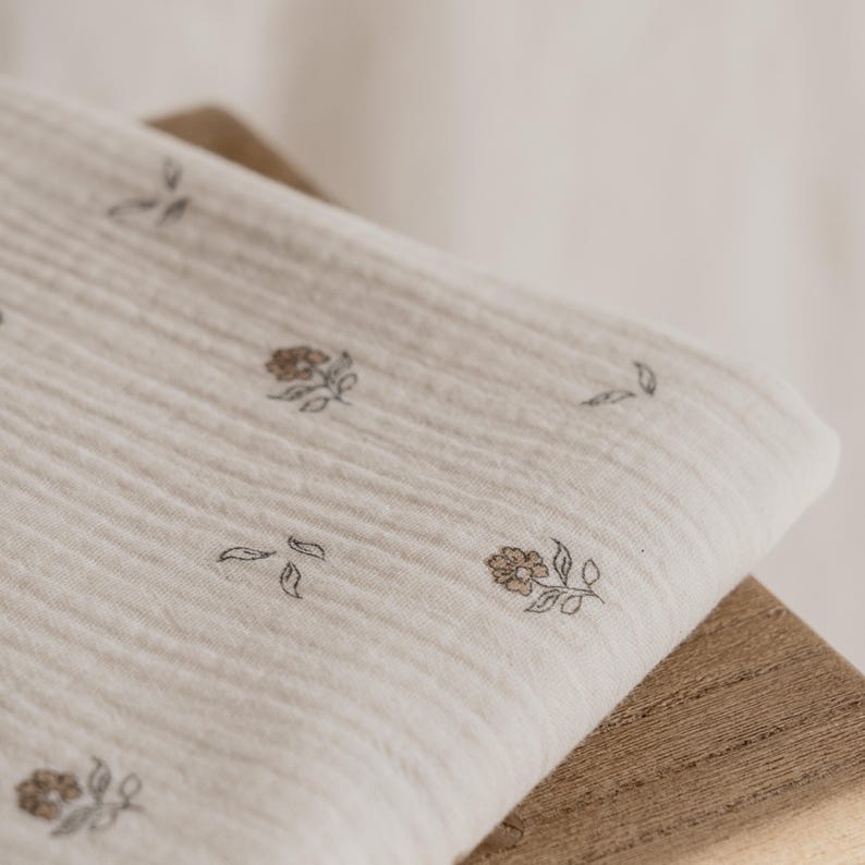 May include: A folded, off-white muslin blanket with a floral pattern. The flowers are a muted brown color and are scattered across the fabric. The blanket is resting on a wooden surface.