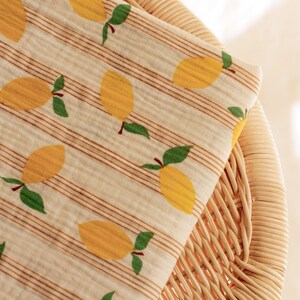 May include: A folded fabric with a repeating pattern of yellow lemons with green leaves and brown stems. The fabric has a cream background with thin brown stripes. The fabric is resting on a woven wicker chair.