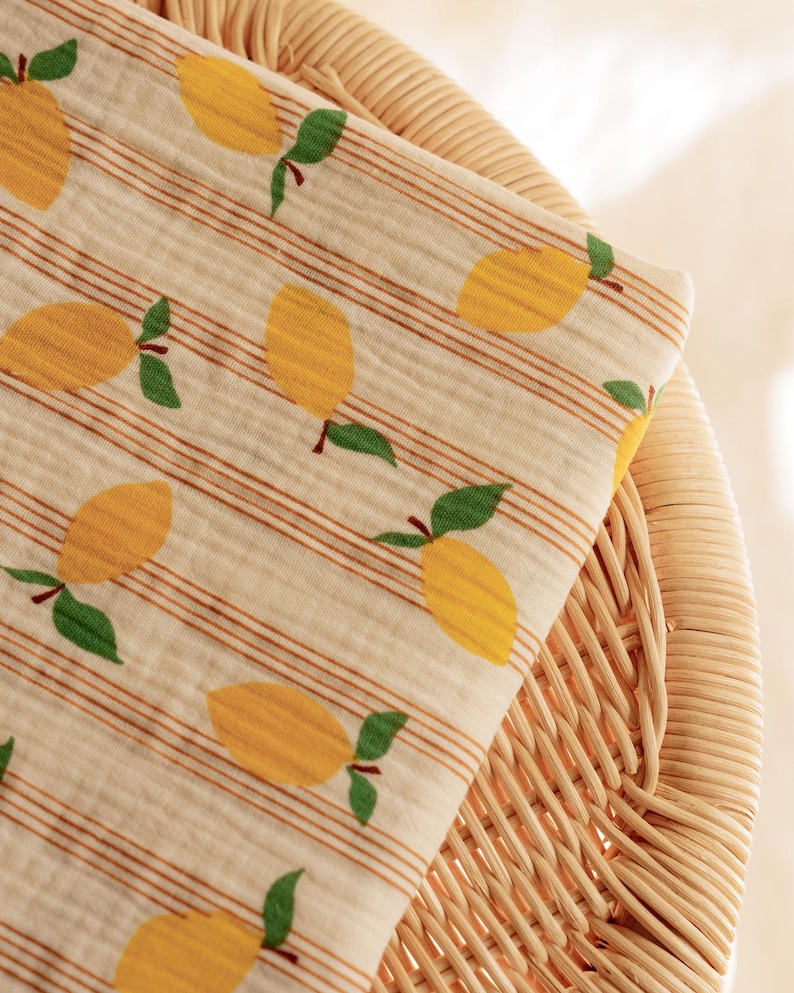 May include: A folded fabric with a repeating pattern of yellow lemons with green leaves and brown stems. The fabric has a cream background with thin brown stripes. The fabric is resting on a woven wicker chair.