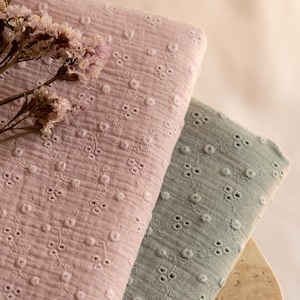 以下が含まれることがあります： 2 つの折り畳まれた生地のクローズアップ。1 つは花の刺繍模様のソフトピンク、もう 1 つは落ち着いたグリーンです。生地は質感があり、わずかに縮んだ外観をしています。ドライフラワーがピンクの生地の上に置かれています。