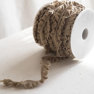 May include: A spool of brown ruffled trim with a white cardboard core. The trim is made of a soft, textured fabric and has a delicate, ruffled edge.