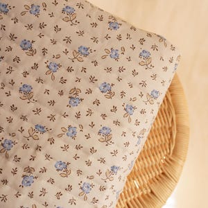 May include: A quilted fabric with a floral pattern of small blue flowers and brown leaves on a cream background. The fabric is folded over a wicker chair, showing the texture and design of the material. The quilting creates a subtle grid pattern.