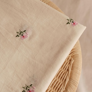 K&ouml;nnte beinhalten: Ein St&uuml;ck cremefarbener Stoff mit einem sich wiederholenden Muster aus kleinen, gestickten rosa Blumen mit gr&uuml;nen Stielen und Bl&auml;ttern. Das Textil hat eine weiche, leicht zerknitterte Textur und liegt gefaltet auf einer hellen Rattanfl&auml;che.