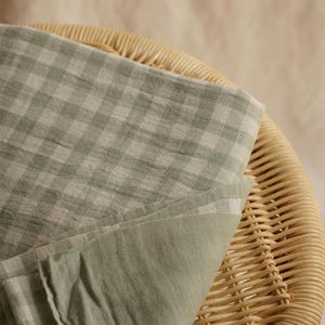May include: A folded piece of light green and white checkered fabric rests on a woven wicker chair. The fabric has a soft, textured appearance, with a solid light green underside. The chair's natural tones provide a neutral backdrop.
