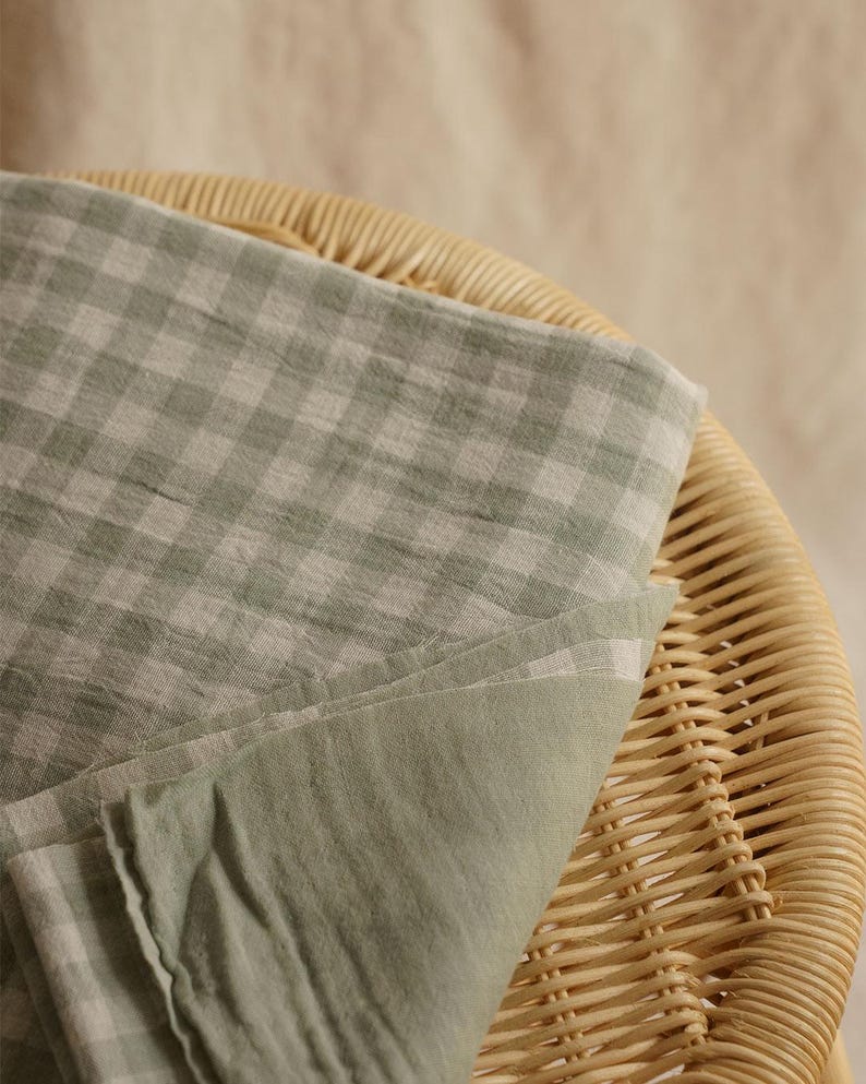 May include: A folded piece of light green and white checkered fabric rests on a woven wicker chair. The fabric has a soft, textured appearance, with a solid light green underside. The chair's natural tones provide a neutral backdrop.