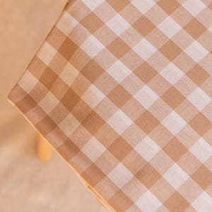Pode incluir: Um tecido quadrado com um padrão clássico de xadrez vichy em tons de bege claro e branco. O material está colocado sobre uma superfície, exibindo um design de grade consistente em toda a área visível do objeto.