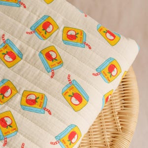 May include: A cream-colored, quilted pillow with a repeating pattern of apple juice boxes. Each box is yellow with a red apple graphic, blue accents, and a red straw. The pillow is on a woven wicker surface.