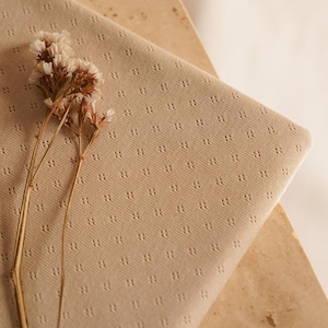 Op de afbeelding: Een close-up van een beige stof met een subtiel, herhalend patroon van kleine stippen. De stof is zacht en textuurrijk en ligt op een lichtbruin oppervlak. Een takje gedroogde witte bloemen ligt op de stof.