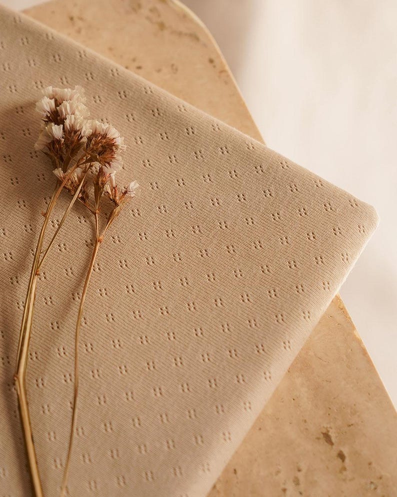 Op de afbeelding: Een close-up van een beige stof met een subtiel, herhalend patroon van kleine stippen. De stof is zacht en textuurrijk en ligt op een lichtbruin oppervlak. Een takje gedroogde witte bloemen ligt op de stof.