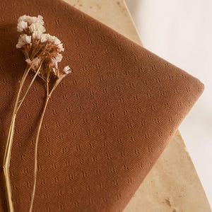 Op de afbeelding: Een close-up van een bruine stof met een textuurpatroon. De stof is gevouwen en heeft een klein boeket witte bloemen erop.