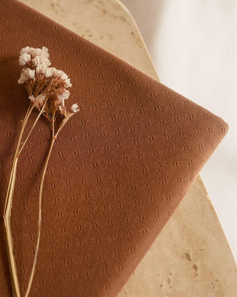 Op de afbeelding: Een close-up van een bruine stof met een textuurpatroon. De stof is gevouwen en heeft een klein boeket witte bloemen erop.