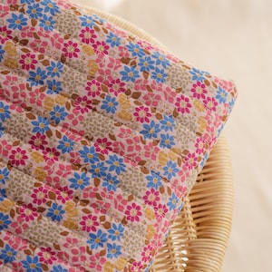 Puede incluir: Primer plano de un cojín con estampado floral. El cojín presenta un patrón repetido de pequeñas flores rosas, azules y amarillas sobre un fondo beige claro. La tela está acolchada con costuras horizontales.