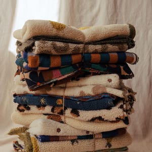 Puede incluir: Una pila de mantas de forro polar sherpa plegadas y atadas con cordel fino. Las mantas presentan varios patrones, incluyendo rayas azules y naranjas, estampados florales oscuros, manchas de leopardo y pequeños motivos de sol.
