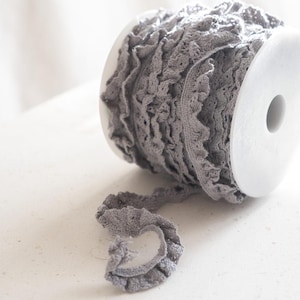 May include: A spool of gray ruffled trim lace. The lace is delicate and has a soft, romantic look.