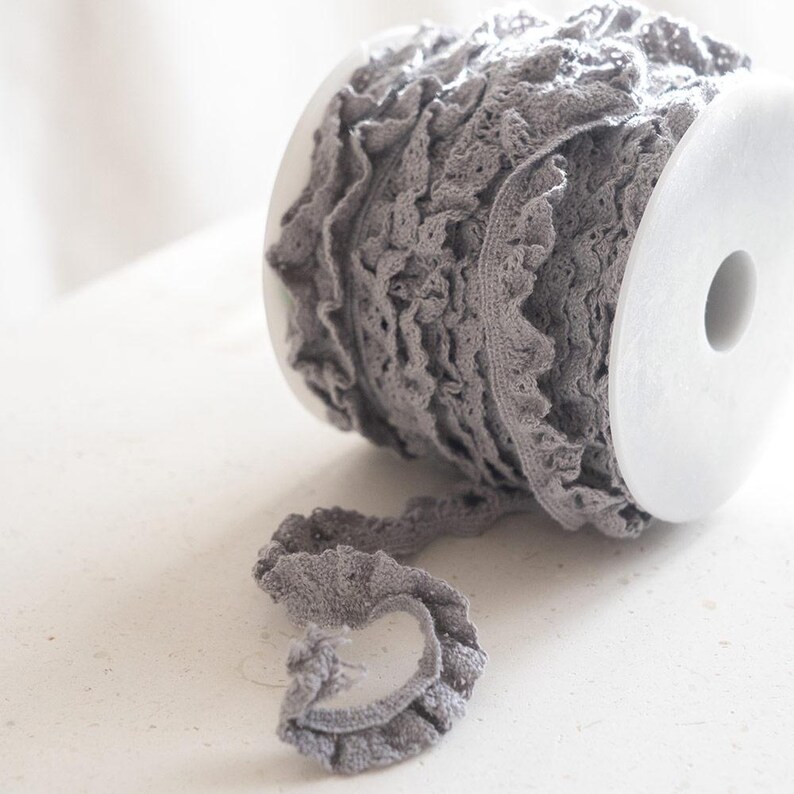 May include: A spool of gray ruffled trim lace. The lace is delicate and has a soft, romantic look.