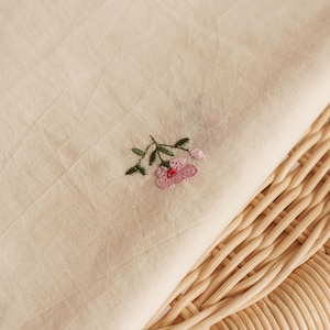K&ouml;nnte beinhalten: Ein St&uuml;ck hellbeiger Stoff mit einer kleinen, feinen Stickerei einer rosa Blume mit gr&uuml;nen Bl&auml;ttern und Stielen. Der Stoff hat eine sichtbare Textur und liegt auf einer hellen, geflochtenen Oberfl&auml;che mit einem abgerundeten Muster.