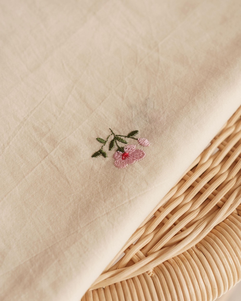 K&ouml;nnte beinhalten: Ein St&uuml;ck hellbeiger Stoff mit einer kleinen, feinen Stickerei einer rosa Blume mit gr&uuml;nen Bl&auml;ttern und Stielen. Der Stoff hat eine sichtbare Textur und liegt auf einer hellen, geflochtenen Oberfl&auml;che mit einem abgerundeten Muster.