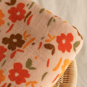 Könnte beinhalten: Eine gefaltete, pfirsichfarbene Fleece-Decke mit einem sich wiederholenden Blumenmuster in Orange, Braun und Gelb. Die Decke liegt auf einem Korbstuhl und zeigt ihre weiche Textur und ihr Design.