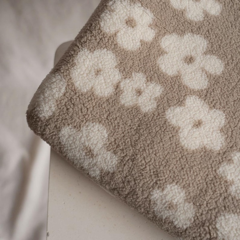 Puede incluir: Primer plano de una tela beige con un patr&oacute;n floral blanco. La tela tiene una textura suave y peluda.