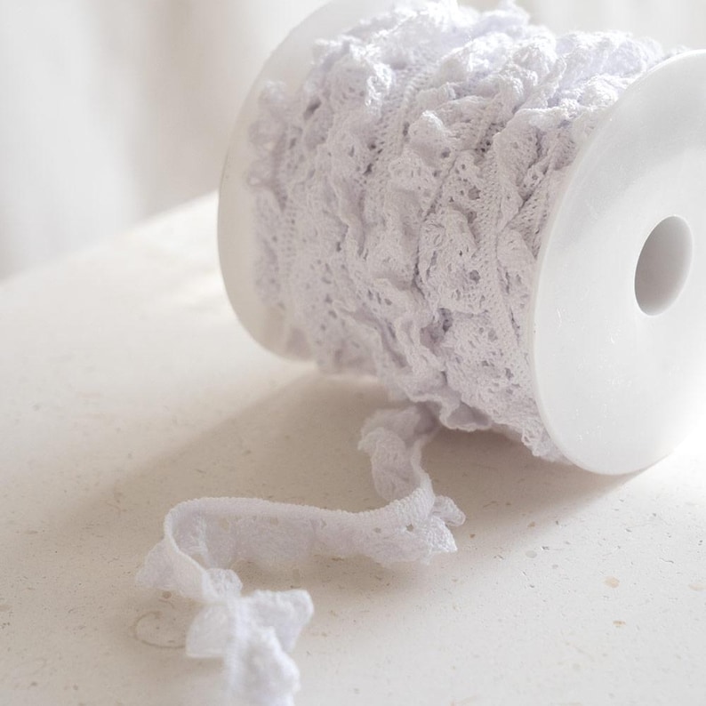 May include: A spool of white lace trim with a ruffled edge. The lace is wound around a white plastic spool.