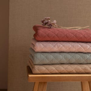 Könnte beinhalten: Ein Stapel von vier gesteppten Decken in verschiedenen Schattierungen von Rosa, Blau und Braun. Die Decken sind gefaltet und auf einem Holzhocker gestapelt.