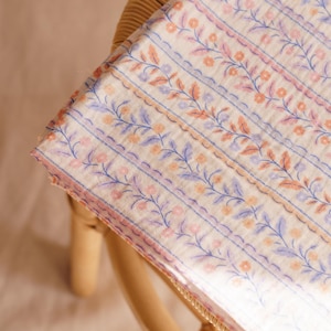 May include: A folded fabric with a floral pattern. The design features rows of blue, pink, and peach-colored flowers and leaves on a cream background. The fabric is resting on a light brown wicker chair.