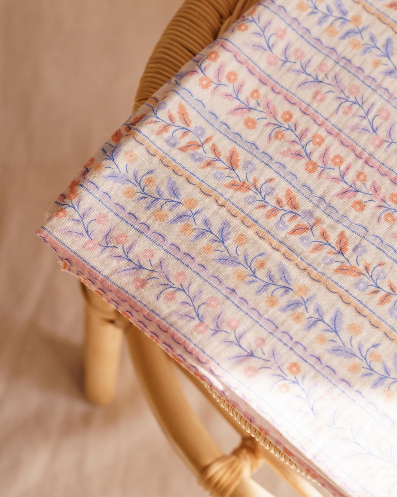 May include: A folded fabric with a floral pattern. The design features rows of blue, pink, and peach-colored flowers and leaves on a cream background. The fabric is resting on a light brown wicker chair.