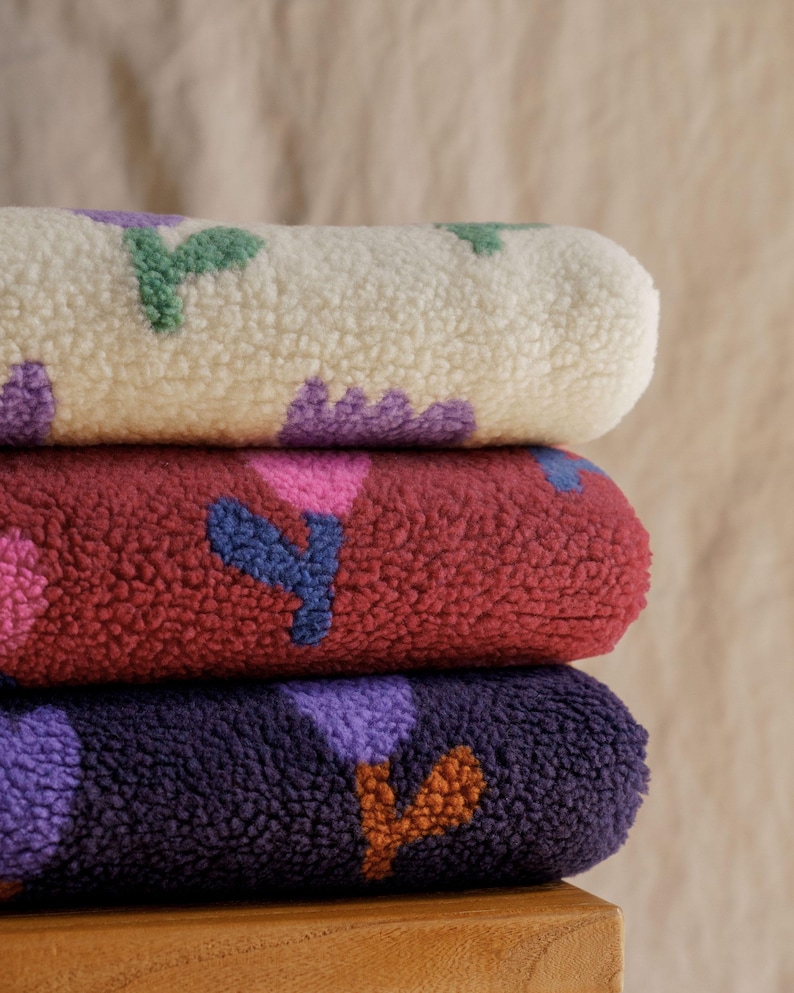 May include: Three rolled fleece blankets stacked on top of each other. The top blanket is cream-colored with green and purple floral patterns. The middle blanket is burgundy with pink and blue floral patterns. The bottom blanket is dark purple with orange and purple floral patterns.