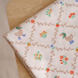 Puede incluir: Tela color crema con un patrón repetido de patos, flores y bayas. Los patos llevan sombreros y botas, y el diseño incluye elementos florales y de vid. La tela está doblada sobre una superficie de madera clara.