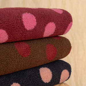 May include: A stack of three folded pieces of textured sherpa fleece fabric. The top piece is deep red with pink polka dots, the middle piece is brown with red polka dots, and the bottom piece is dark charcoal gray with light pink polka dots.