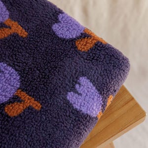 May include: Close-up of a plush, textured fabric in deep navy blue, adorned with a pattern of purple and orange shapes. The fabric is folded over a wooden surface, showcasing its soft texture and color contrast.
