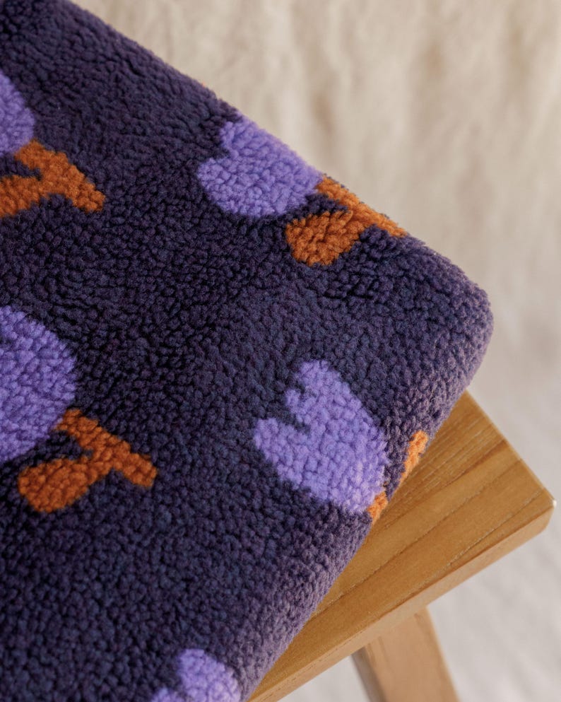 May include: Close-up of a plush, textured fabric in deep navy blue, adorned with a pattern of purple and orange shapes. The fabric is folded over a wooden surface, showcasing its soft texture and color contrast.