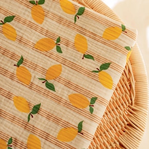 May include: A folded, cream-colored fabric with a repeating pattern of yellow lemons and green leaves. The fabric has thin, brown horizontal stripes. The fabric is resting on a woven wicker surface.