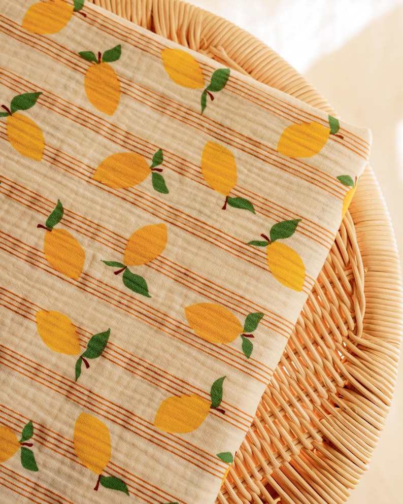 May include: A folded, cream-colored fabric with a repeating pattern of yellow lemons and green leaves. The fabric has thin, brown horizontal stripes. The fabric is resting on a woven wicker surface.