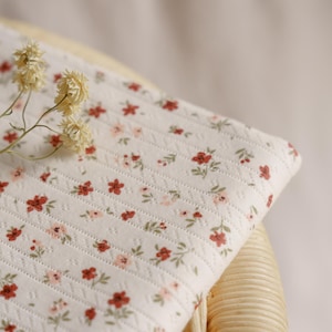 May include: A folded piece of white fabric with a floral pattern of small red and pink flowers and green leaves. The fabric is placed on a woven wicker chair. Dried flowers are placed on the fabric.
