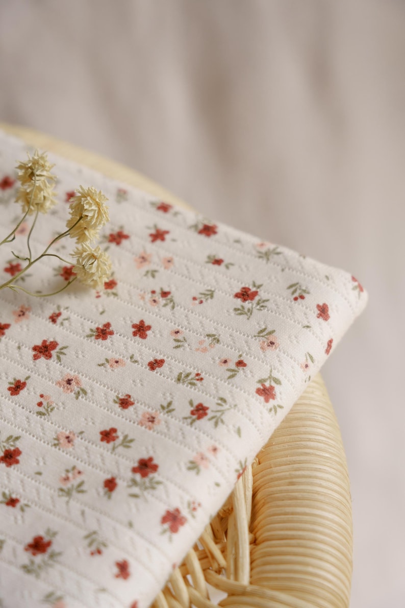 May include: A folded piece of white fabric with a floral pattern of small red and pink flowers and green leaves. The fabric is placed on a woven wicker chair. Dried flowers are placed on the fabric.