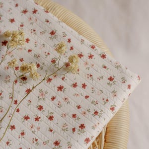 May include: A white, quilted fabric with a floral pattern of small red and pink flowers. The fabric is draped over a woven wicker chair, with dried flowers in the foreground.