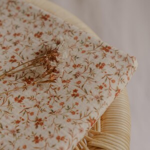 May include: A folded piece of fabric with a delicate floral pattern in shades of orange, pink, and green on a cream background. The fabric is resting on a woven chair, with dried flowers on top.