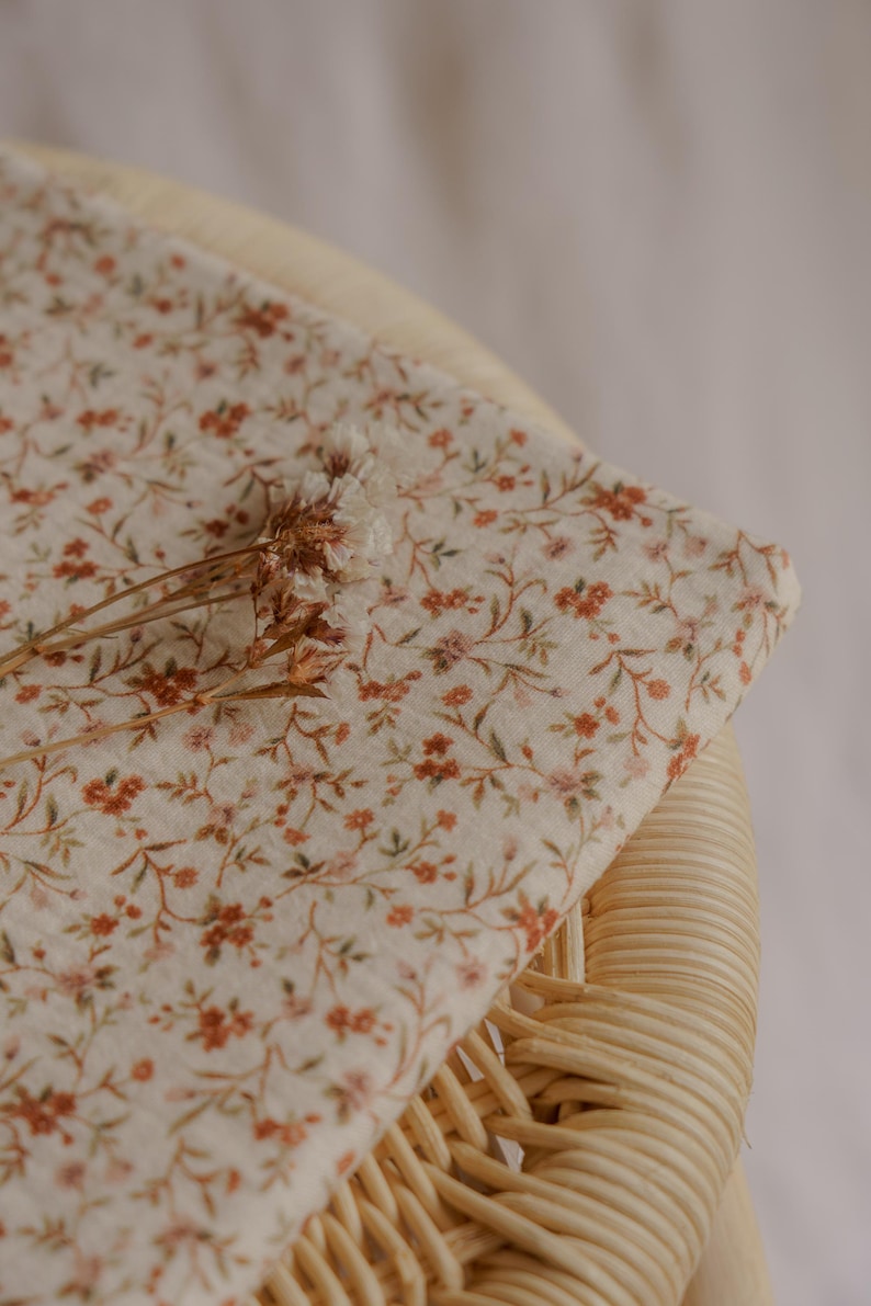 May include: A folded piece of fabric with a delicate floral pattern in shades of orange, pink, and green on a cream background. The fabric is resting on a woven chair, with dried flowers on top.