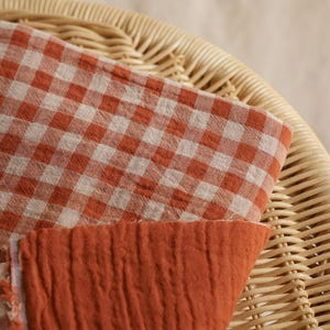 May include: Close-up of two fabric swatches. One is a gingham pattern in orange and white, the other is a solid orange, textured fabric. The fabrics are resting on a woven wicker chair.