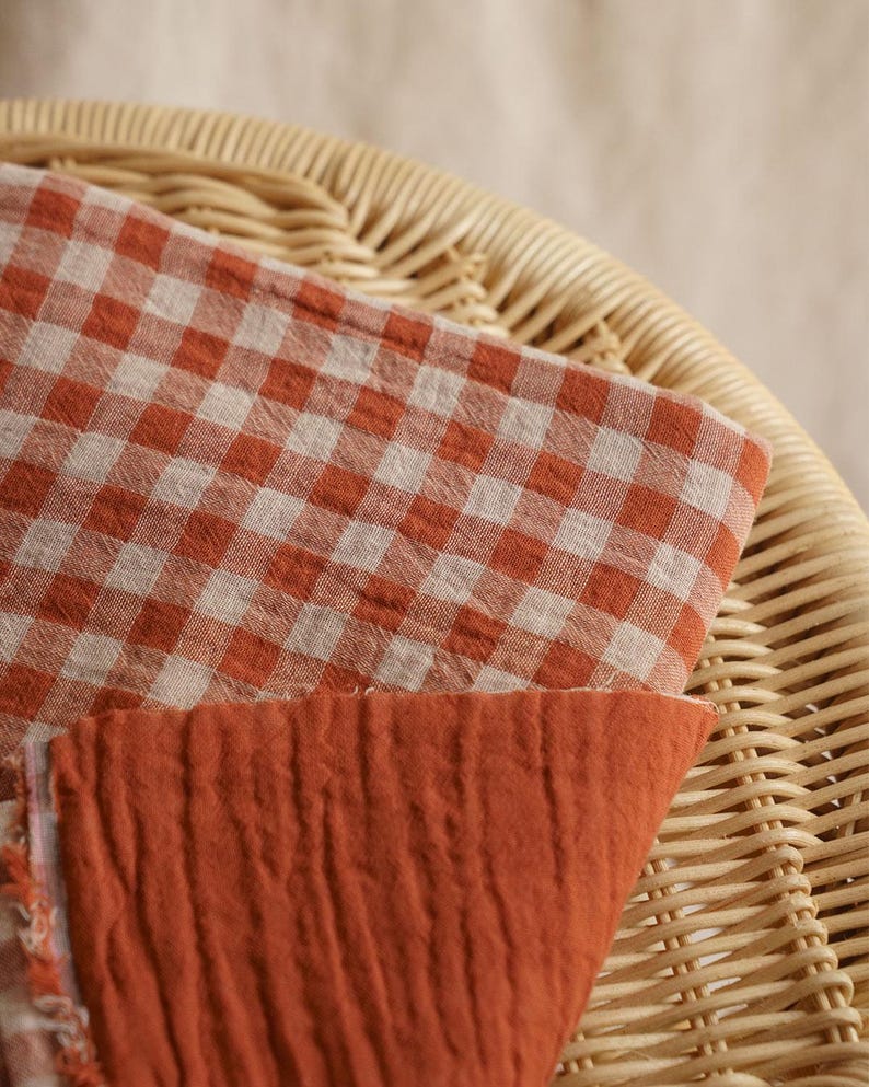 May include: Close-up of two fabric swatches. One is a gingham pattern in orange and white, the other is a solid orange, textured fabric. The fabrics are resting on a woven wicker chair.