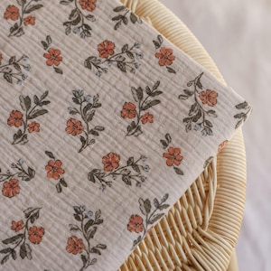 May include: A folded fabric featuring a floral design. The fabric is a pale beige, adorned with a repeating pattern of orange flowers, green leaves, and small blue flowers. It rests on a wicker chair.