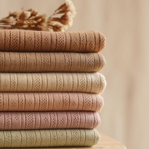 May include: A stack of six folded textiles in varying shades of brown, beige, pink, and green. Each textile has a textured, patterned design. A dried floral arrangement rests on top of the stack.