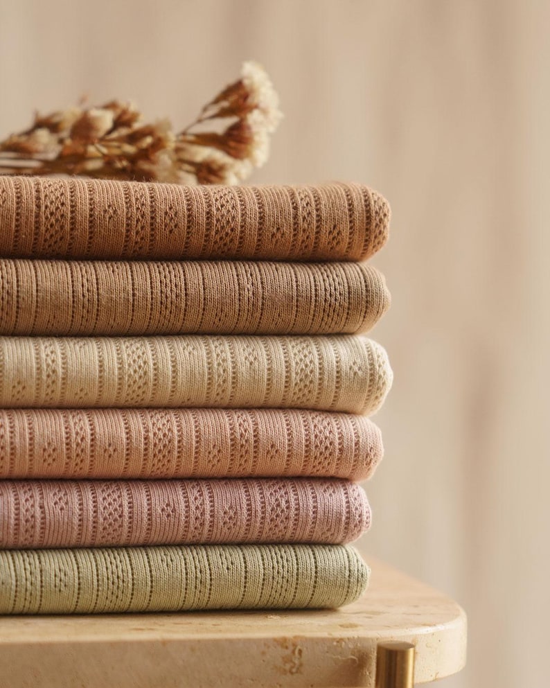 May include: A stack of six folded textiles in varying shades of brown, beige, pink, and green. Each textile has a textured, patterned design. A dried floral arrangement rests on top of the stack.